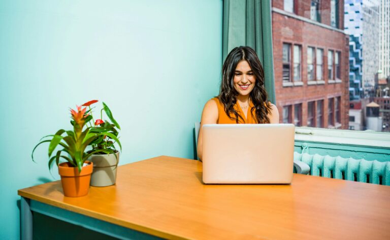 Femme confiante travaillant sur un ordinateur portable dans un bureau lumineux, entourée de paysages urbains et de verdure.